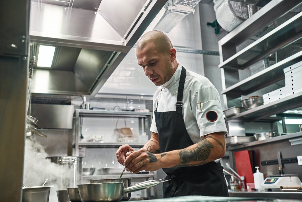 Important moment. Serious professional chef in apron, with several tattoos on his hands cooking in a