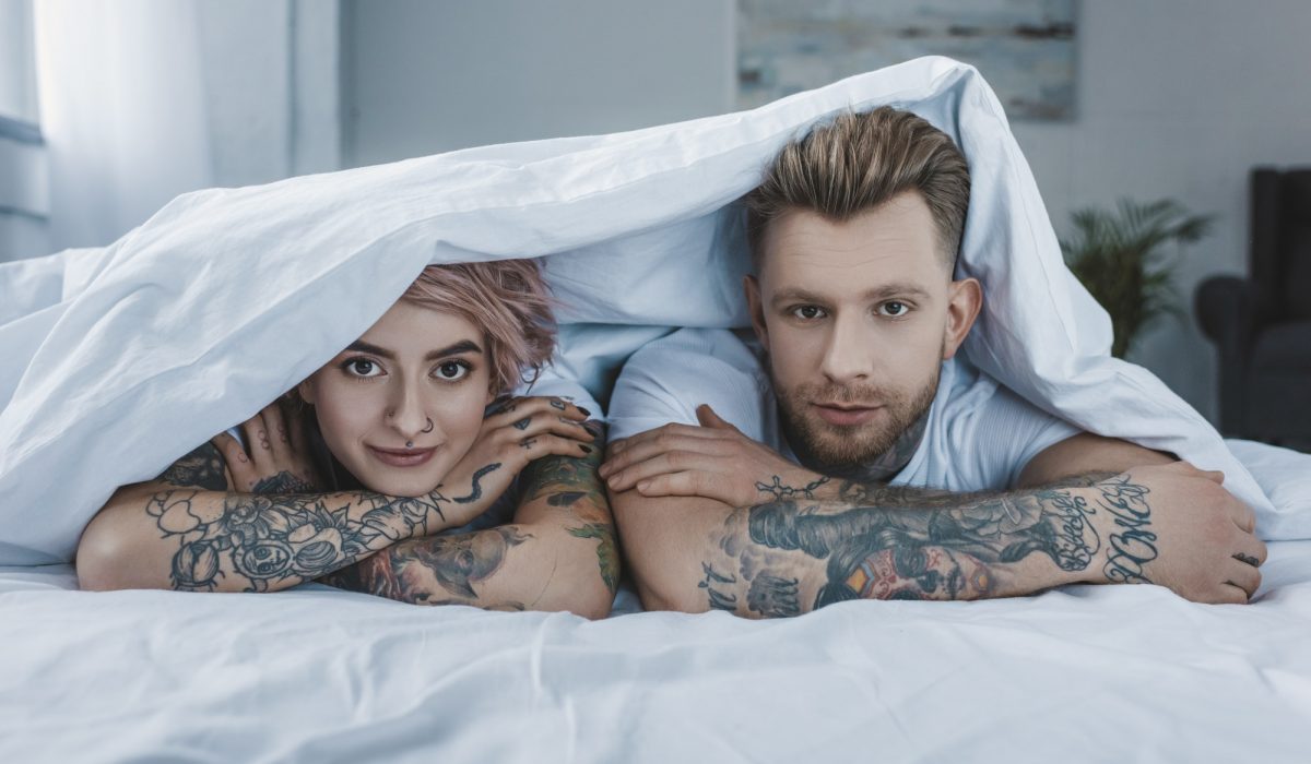 young tattooed couple lying in bed under white blanket